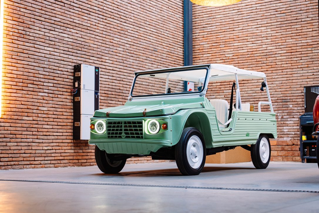 Sage green Méhari inside the Dania Beach atelier with battery modules visible on the brick wall behind